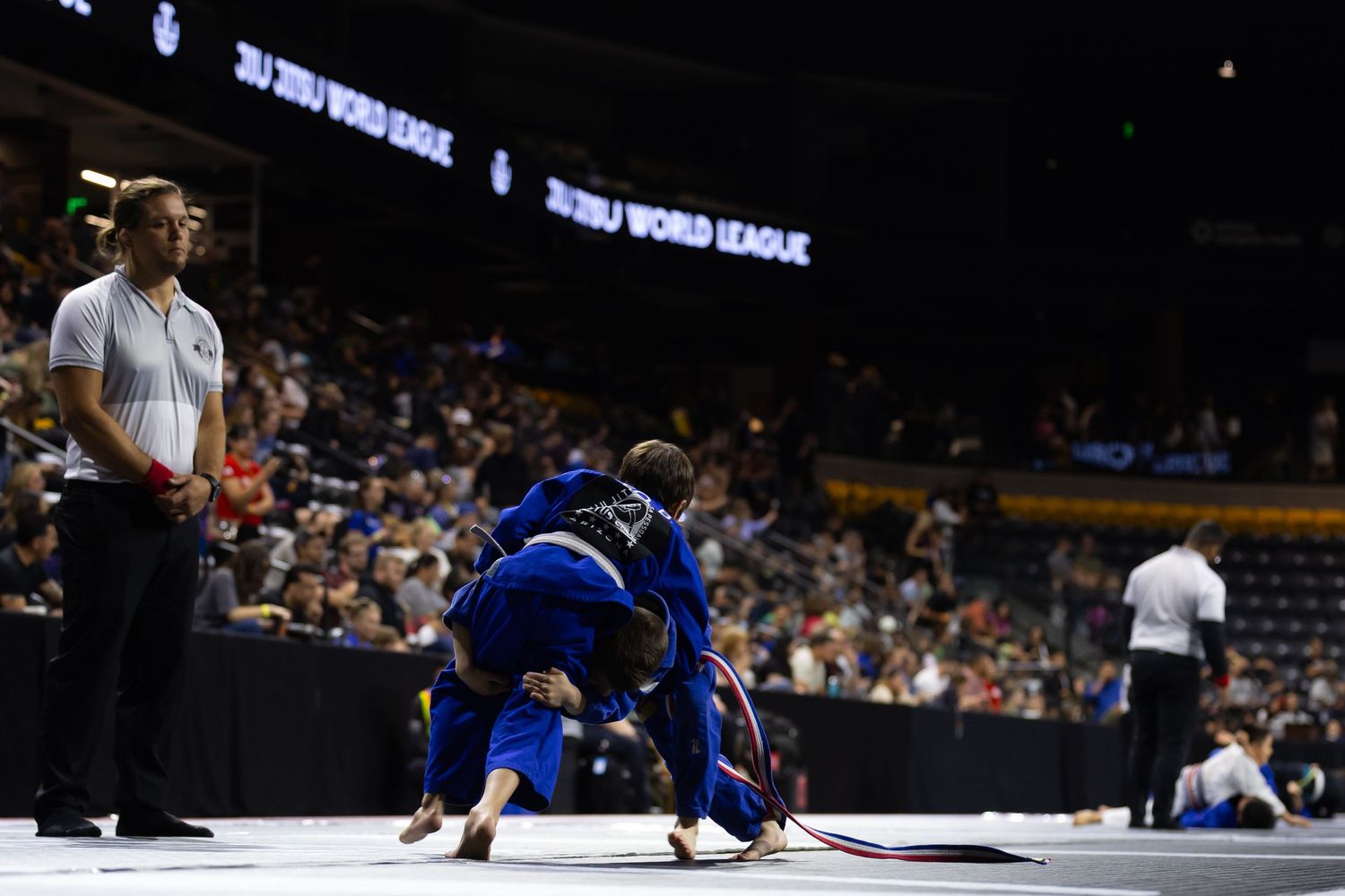 Two wrestlers compete on a mat during a match while a referee observes from the side in an indoor arena.