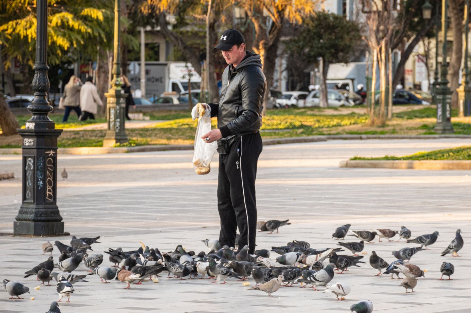A person stands on a paved walkway surrounded by feeding pigeons in an urban park setting during daytime.