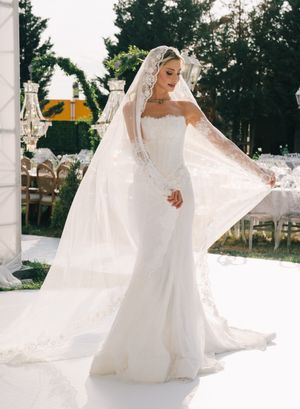 Elegant bride in form-fitting white lace gown and flowing cathedral veil poses outdoors during wedding ceremony.