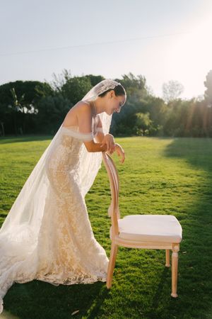A bride in a flowing lace dress leans gracefully against a wooden bench in a sunlit meadow at golden hour.