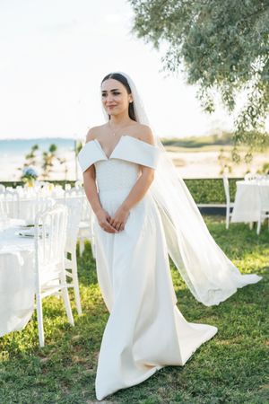 Bride in elegant off-shoulder gown poses on grassy lawn with white chairs in background.