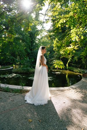 A couple in wedding attire stands by a tranquil pond surrounded by lush green trees on a sunny afternoon.