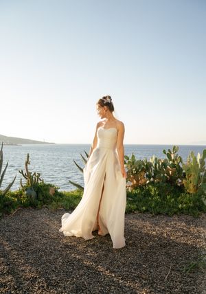 A bride in a flowing white gown walks along a coastal path with ocean views and succulent plants at sunset.