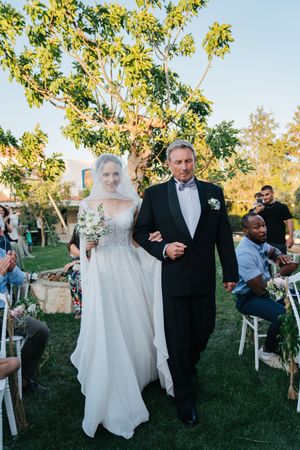 Wedding party processes down outdoor aisle lined with white chairs.