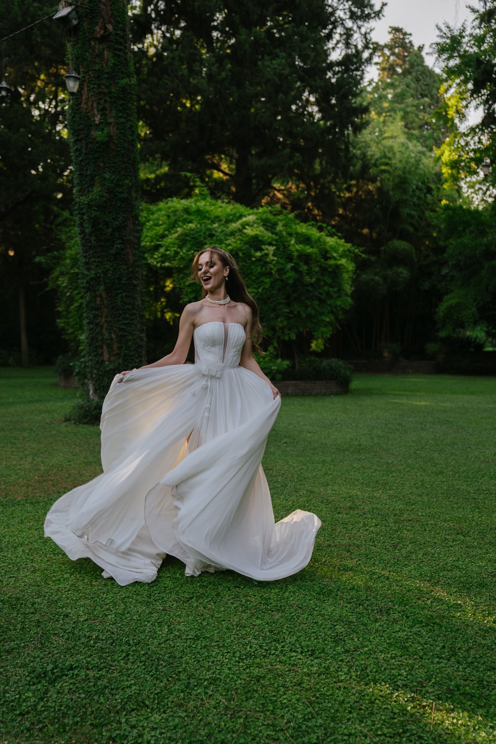 Bride in flowing white dress twirls on lush green lawn during golden hour.