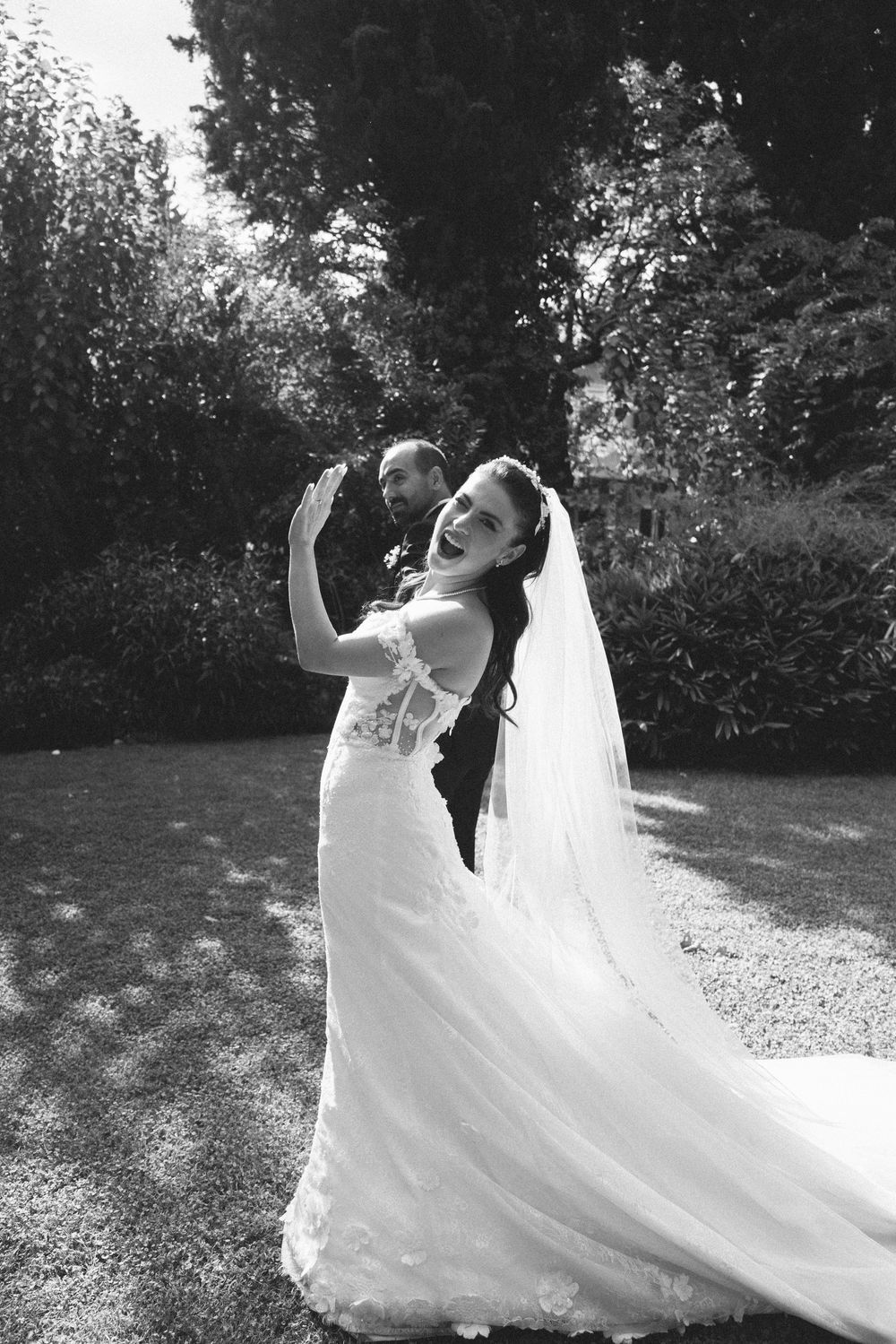 Black and white photo of a bride in a flowing lace gown and veil in a garden setting.