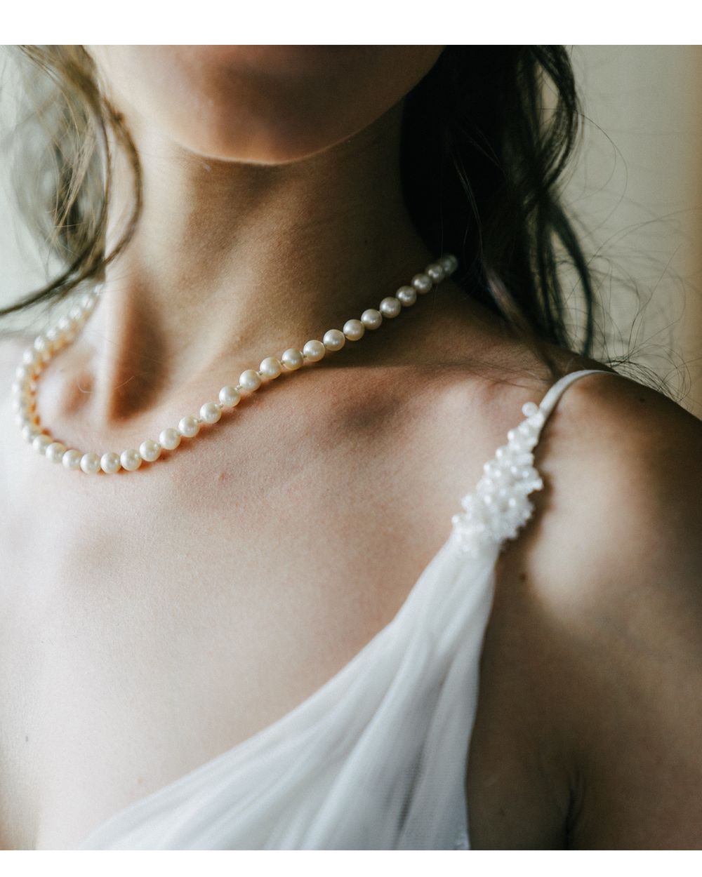 Close-up of an elegant pearl necklace and white dress strap against a soft neutral background.