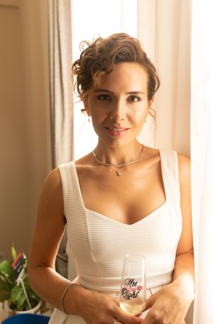 A person in a white sleeveless dress smiles while holding something in their hands near a window with natural light.