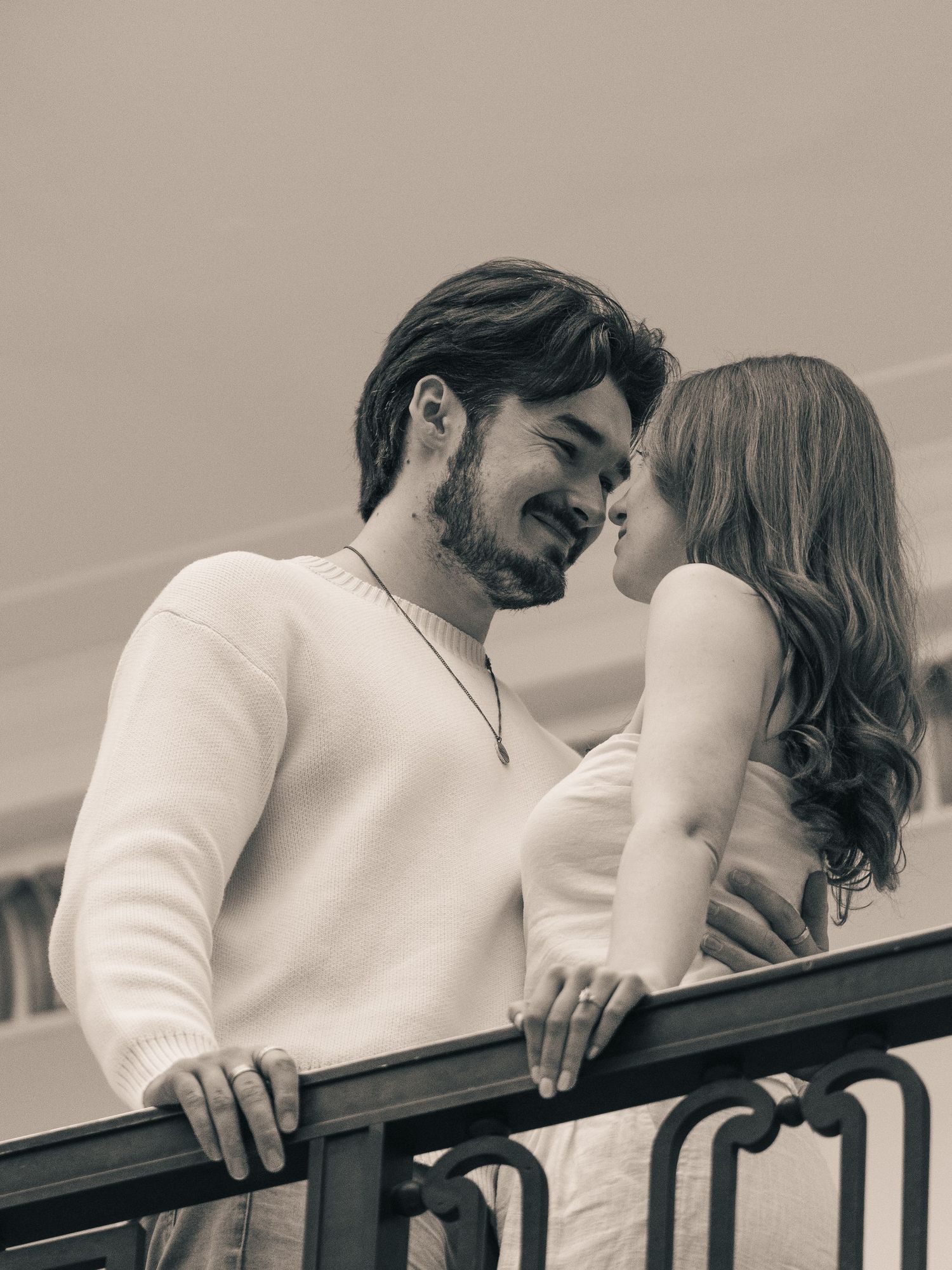 Black and white photo of a bearded man and woman sharing a romantic moment on a balcony railing.