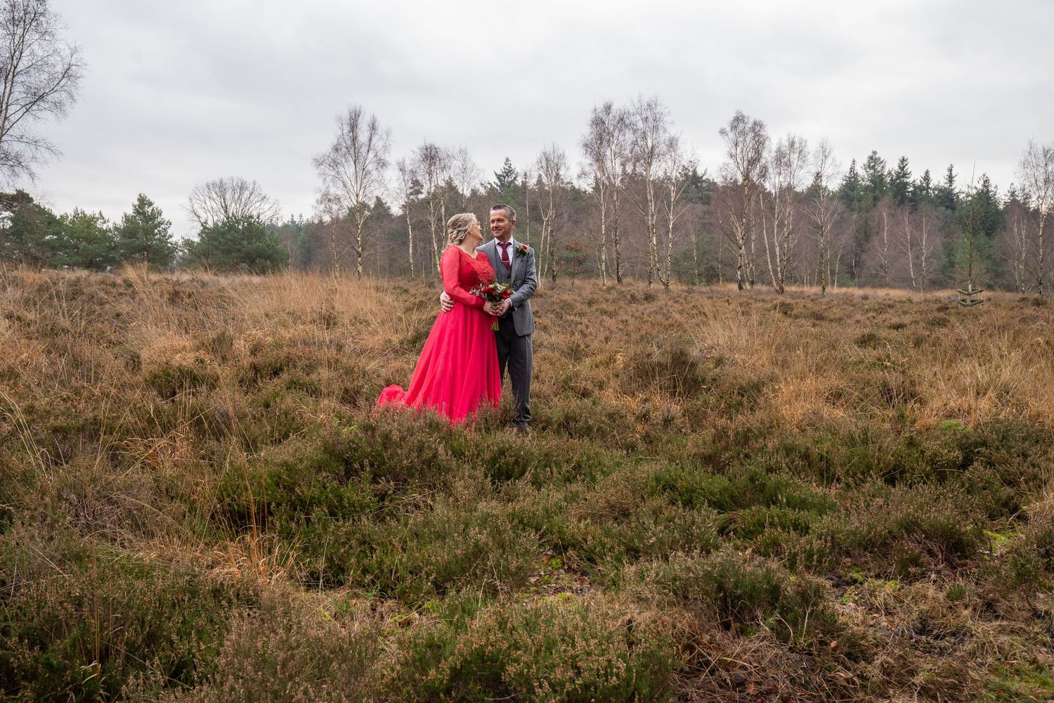 Het bruidspaar poseert in rode en grijze trouwkleding op een uitgestrekt heideveld onder een bewolkte hemel.