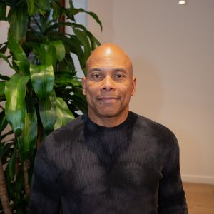 A bald Black man in a dark textured sweater smiles warmly beside a tall indoor plant.