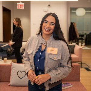 A young woman with long dark hair wearing a gray jacket and name tag smiles at an event.