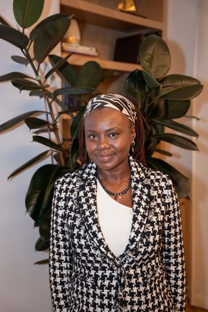 A woman in a houndstooth blazer with a patterned headband smiles beside a large rubber plant.