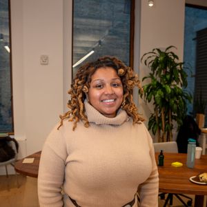 A smiling woman with curly highlighted hair wearing a beige turtleneck sweater in a modern interior.