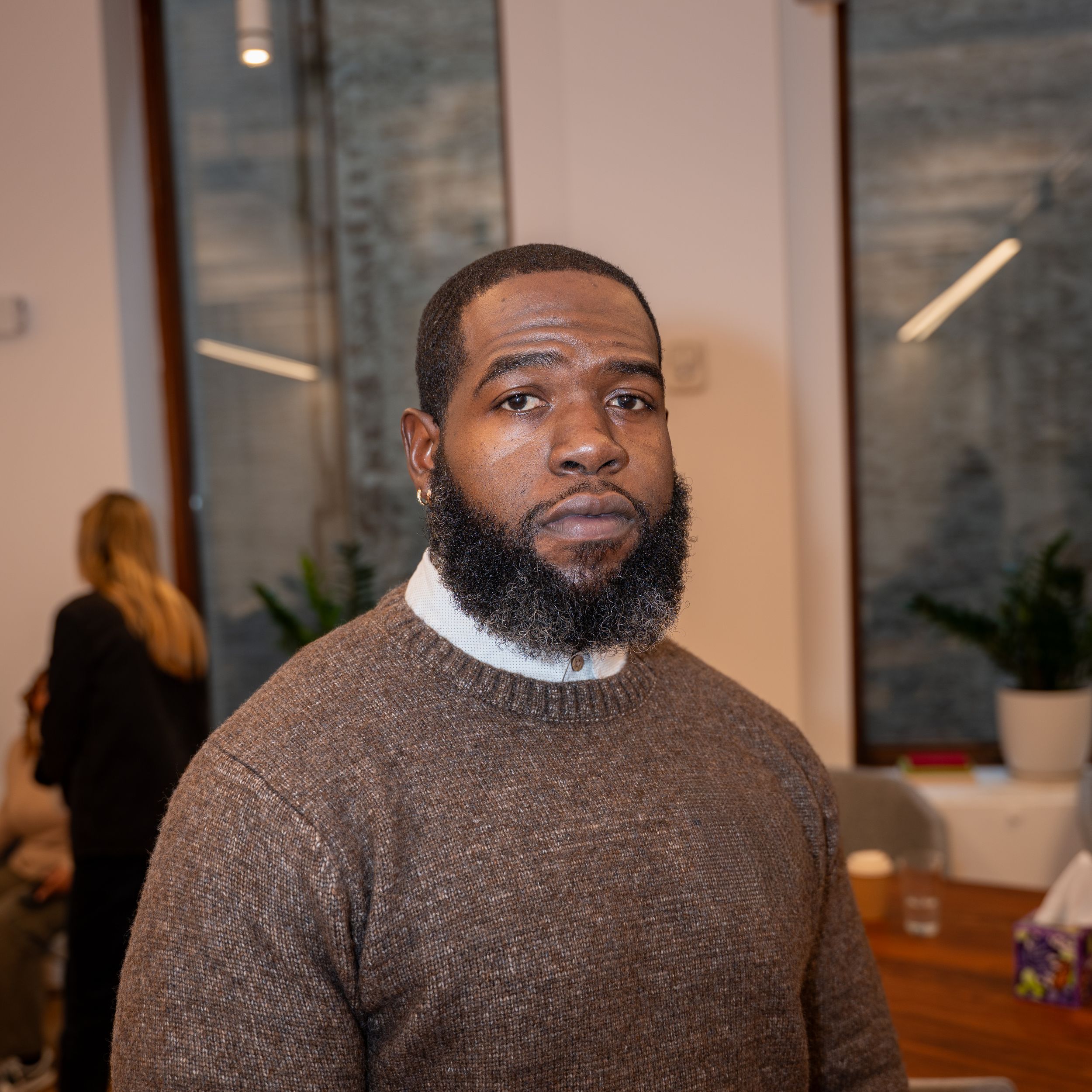 A bearded Black man in a brown sweater poses indoors at a modern office space.