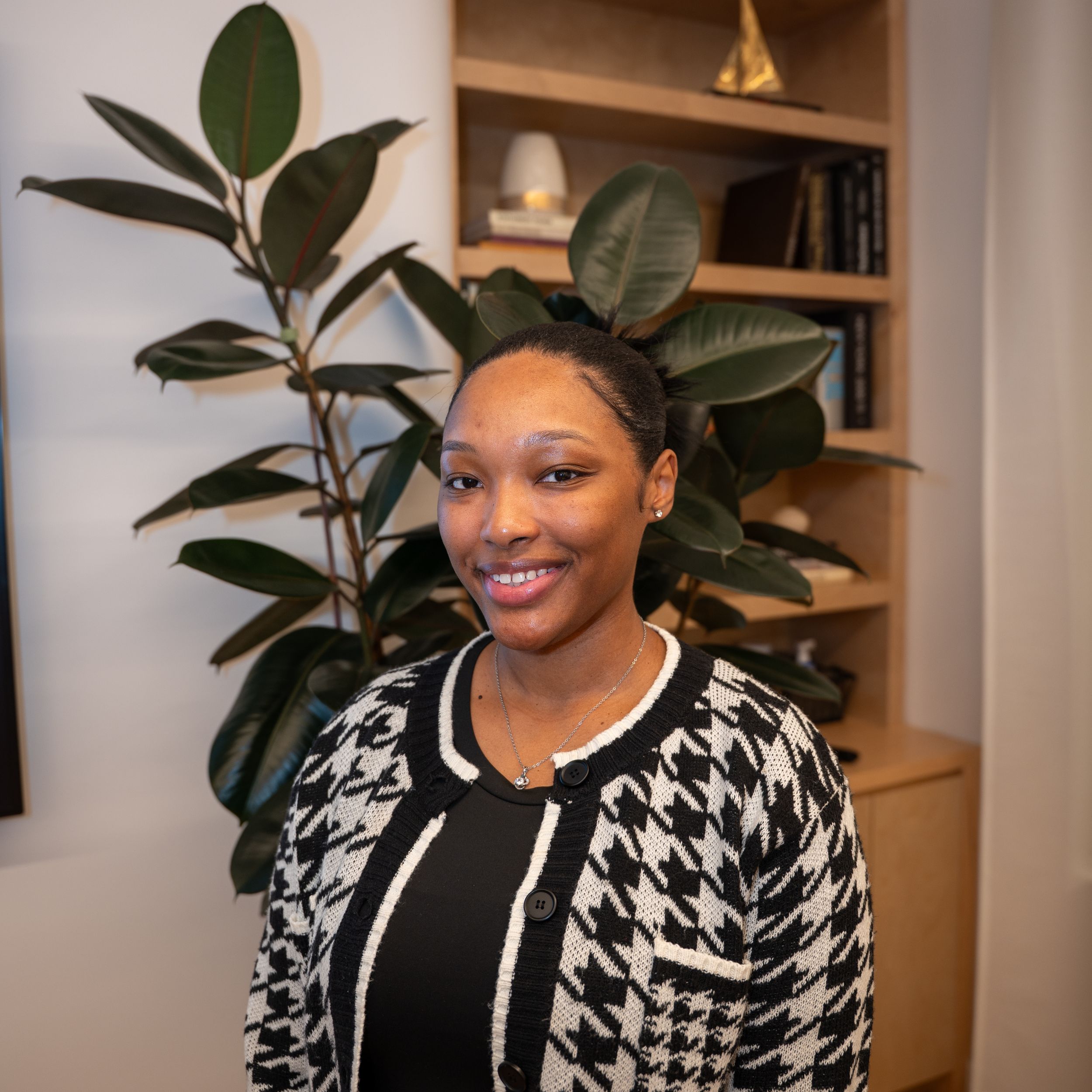 Smiling woman in houndstooth cardigan standing in a modern office with bookshelf and plant.