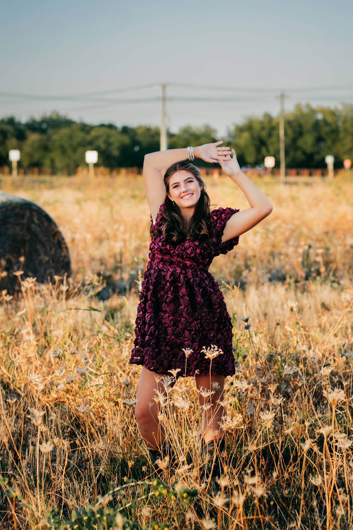 Young woman in dark floral dress posing gracefully in golden field with wildflowers during sunset.