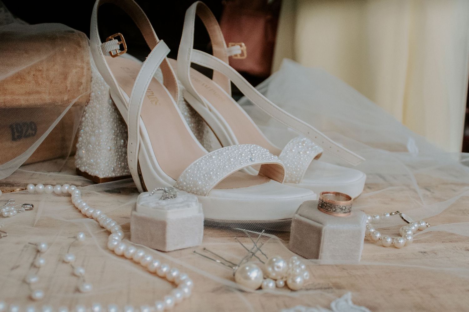 White bridal heels with pearl details displayed alongside gold rings, pearl necklace, and earrings on a wedding day flat lay.