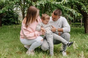 Family of three sitting on green grass under trees, parents smiling at toddler in casual light-colored outfits outdoors.