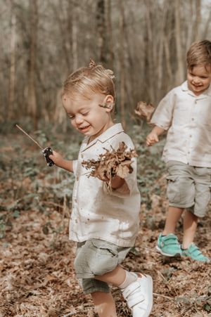 Two young children playing outdoors in a forest, smiling and holding sticks with muddy hands.