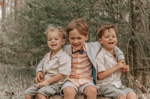 Three young boys in dressy outfits sitting together on a log outdoors, smiling and laughing in a forest setting.