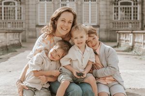 A smiling mother poses outdoors with her three young sons, all dressed in neutral tones, in front of a historic building.