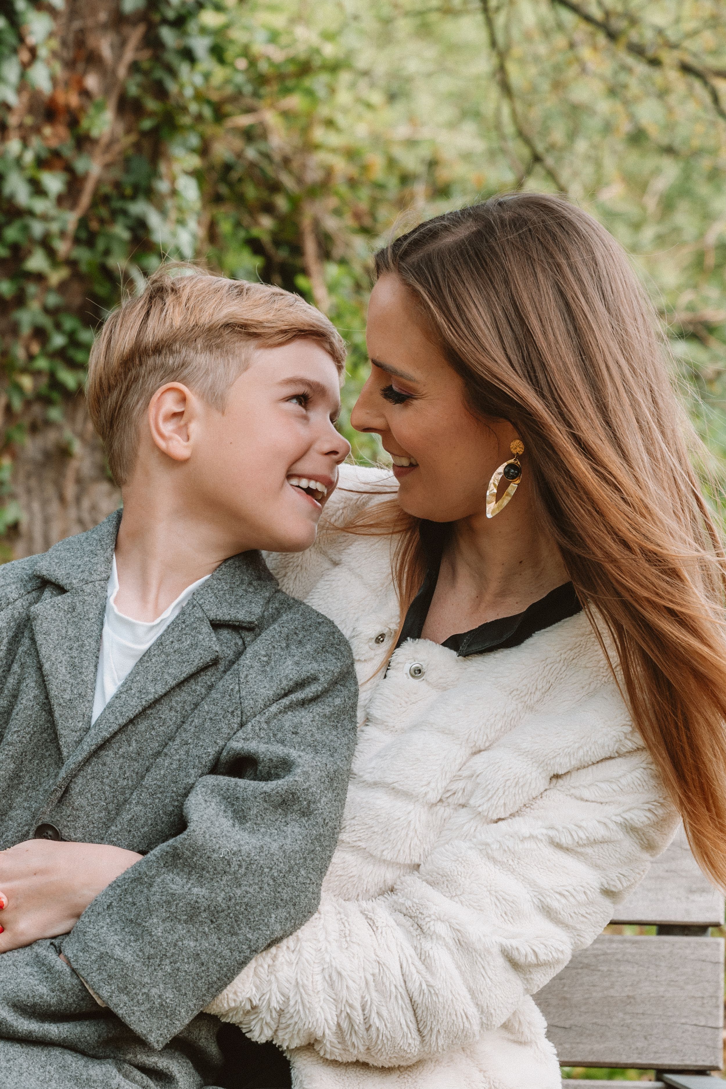 A smiling woman in a white fur coat and her young son in a gray blazer share a joyful moment outdoors.