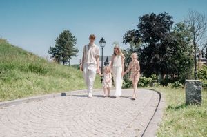 Family of four walking on a cobblestone path in a sunny park, with green trees and blue sky in the background.