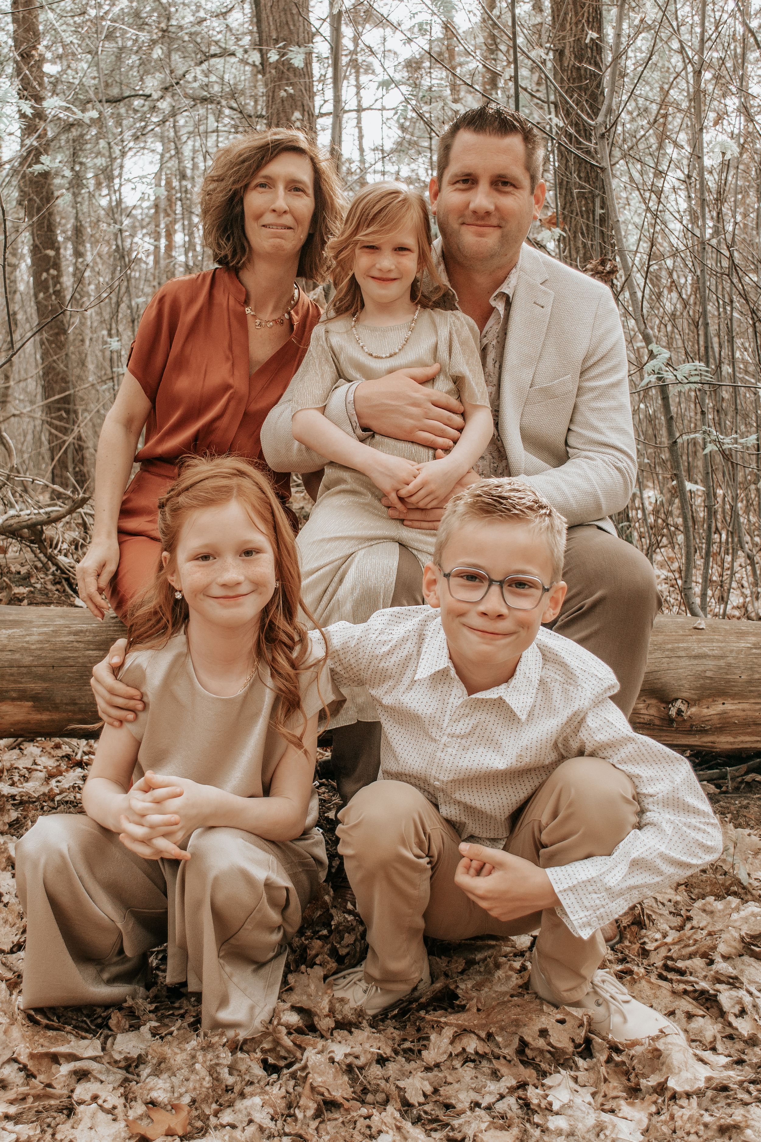 Family of five posing in autumn forest, parents with three children sitting on log among fallen leaves.