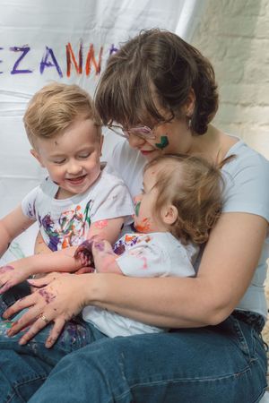 A mother hugs two paint-covered toddlers, smiling joyfully during a colorful finger painting activity.
