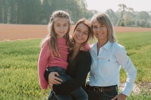 Three generations of women smiling together in a green field, wearing casual clothing on a sunny day.