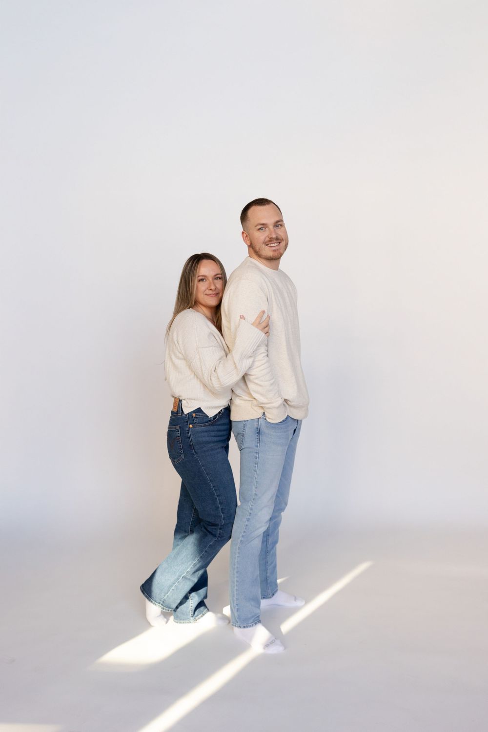 A couple in coordinating cream sweaters and jeans poses playfully against a white background.
