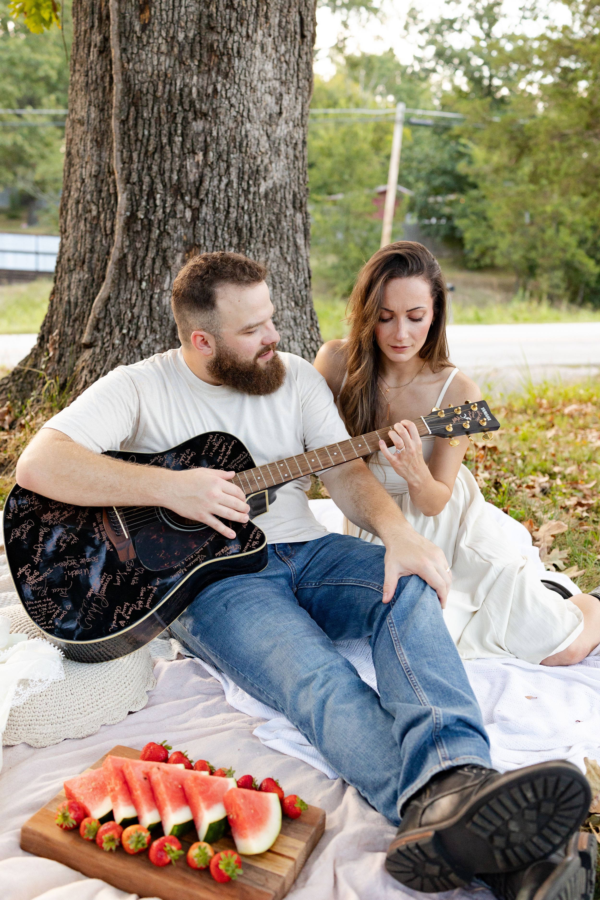 A couple enjoys a romantic picnic with guitar music and fresh fruit under a large tree on a blanket.