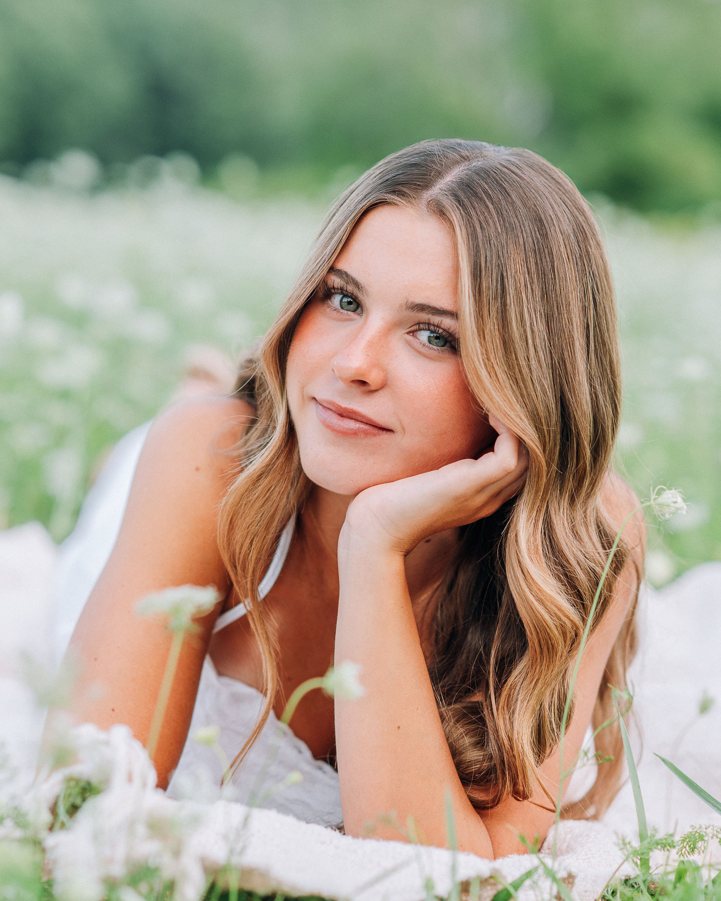Young woman with wavy blonde hair lying in a wildflower field, smiling softly in a white dress.
