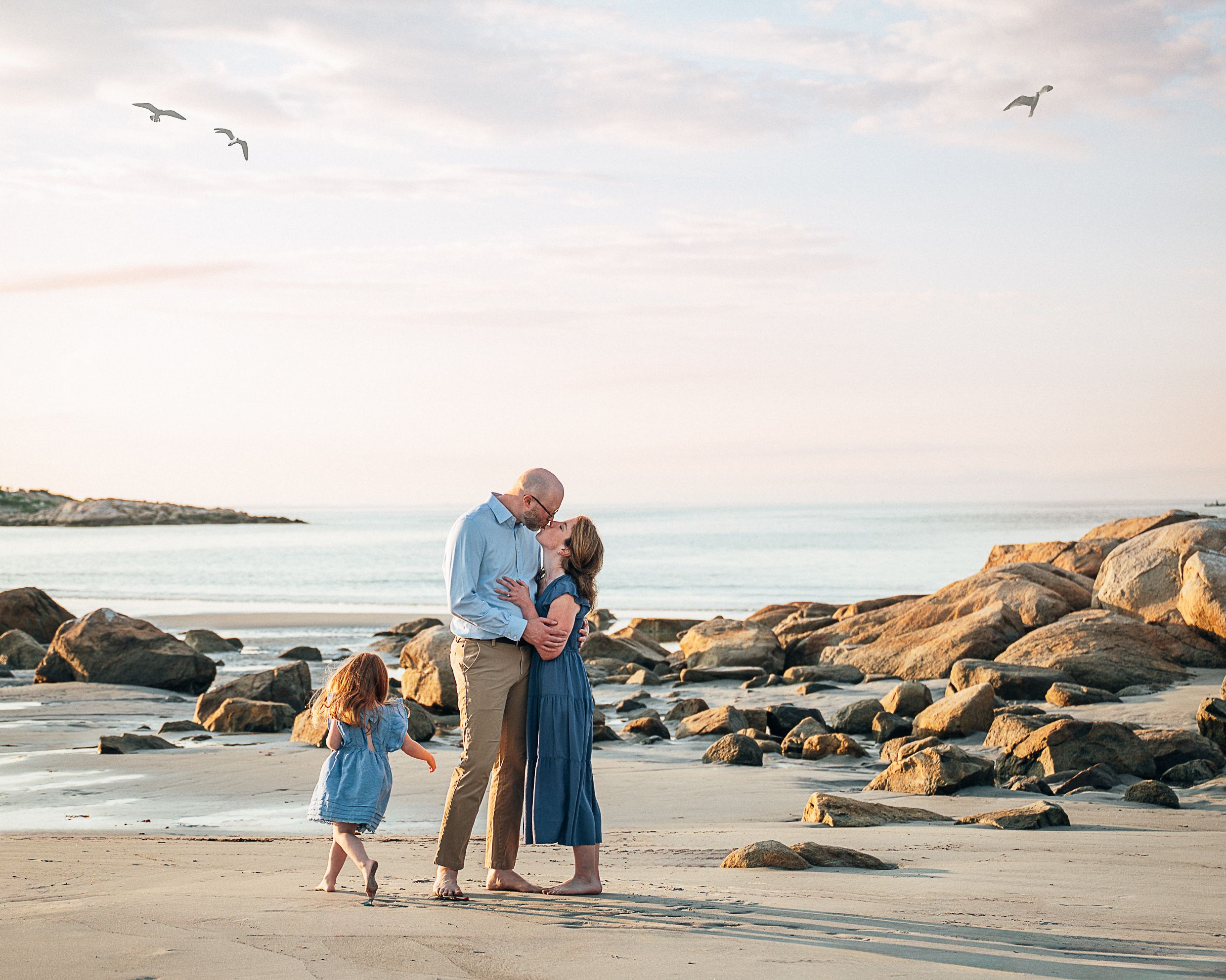 Family of three sharing a tender moment on a rocky beach at sunset with seagulls flying overhead.