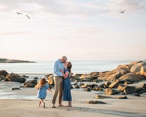 Family of three sharing a tender moment on a rocky beach at sunset with seagulls flying overhead.