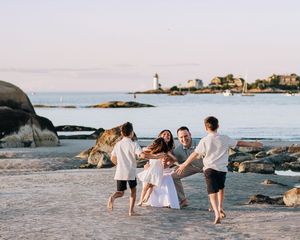 Family playing joyfully on a sandy beach at sunset with a lighthouse and rocky shoreline in the background.