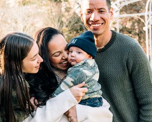 Happy mixed-race family with two women and a man smiling outdoors, holding an adorable baby in a beanie hat.