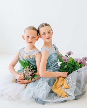 Two young girls in blue ballet dresses holding lilac bouquets pose back-to-back in a bright white studio.
