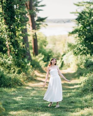 A young woman in a white midi dress walks along a lush green forest path on a sunny day.