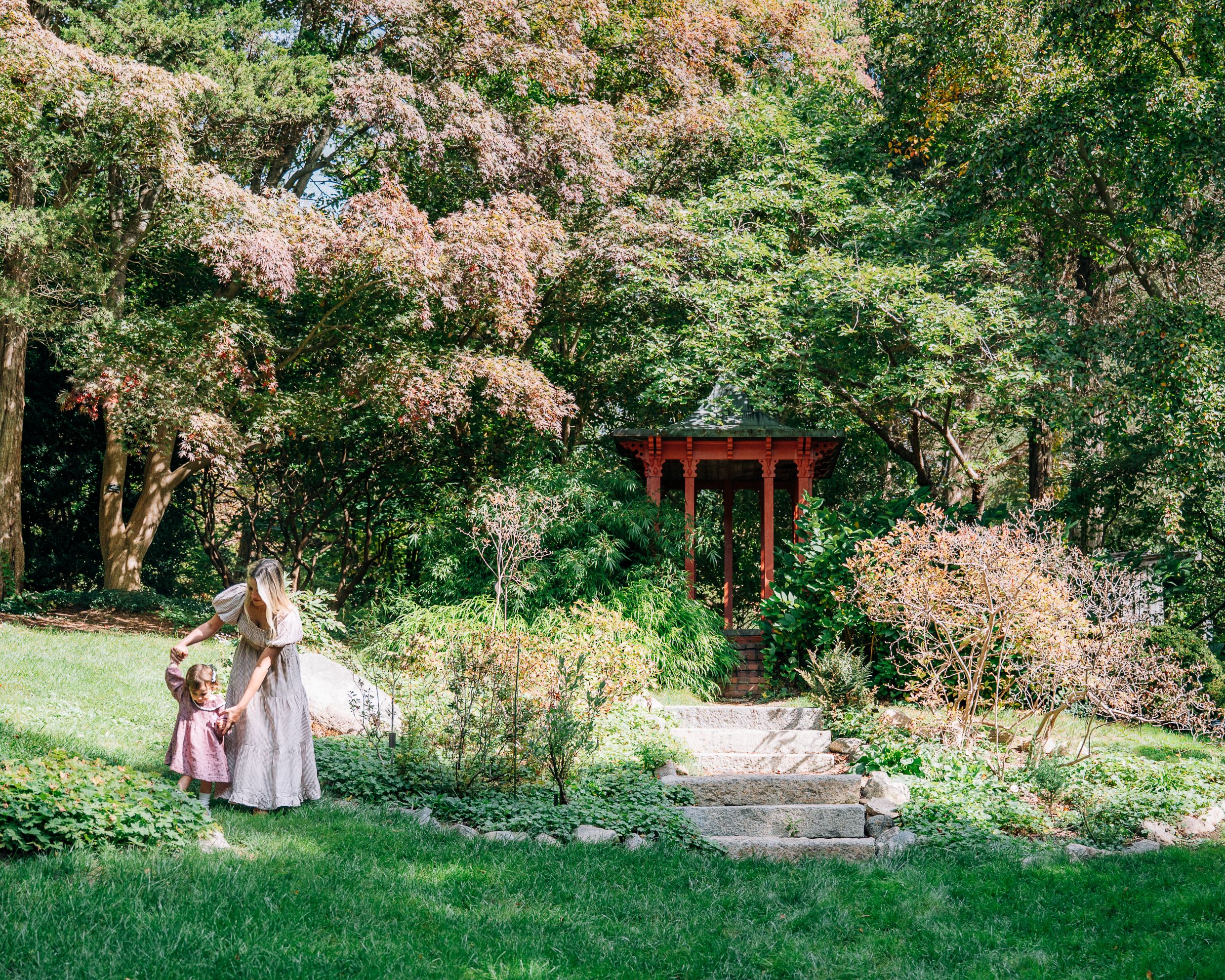 A woman and child walk through a lush garden with a red gazebo and blooming pink trees.