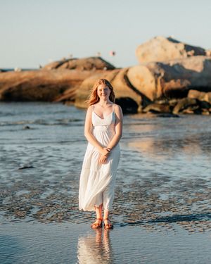 A young woman in a white dress stands barefoot on a reflective beach at golden hour with rocky shores behind her.