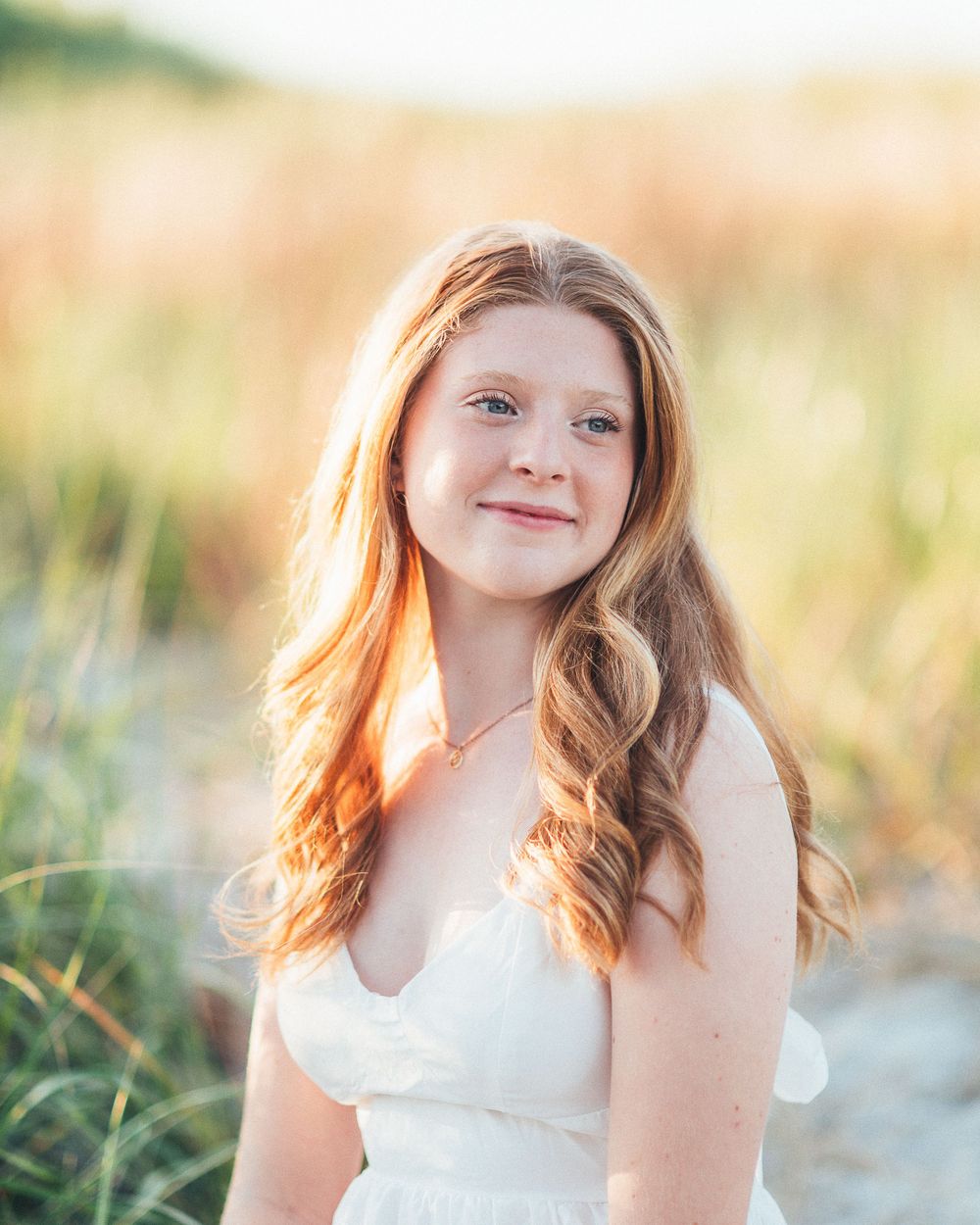 Young girl with long red hair in white dress smiling outdoors at golden hour near beach grass.