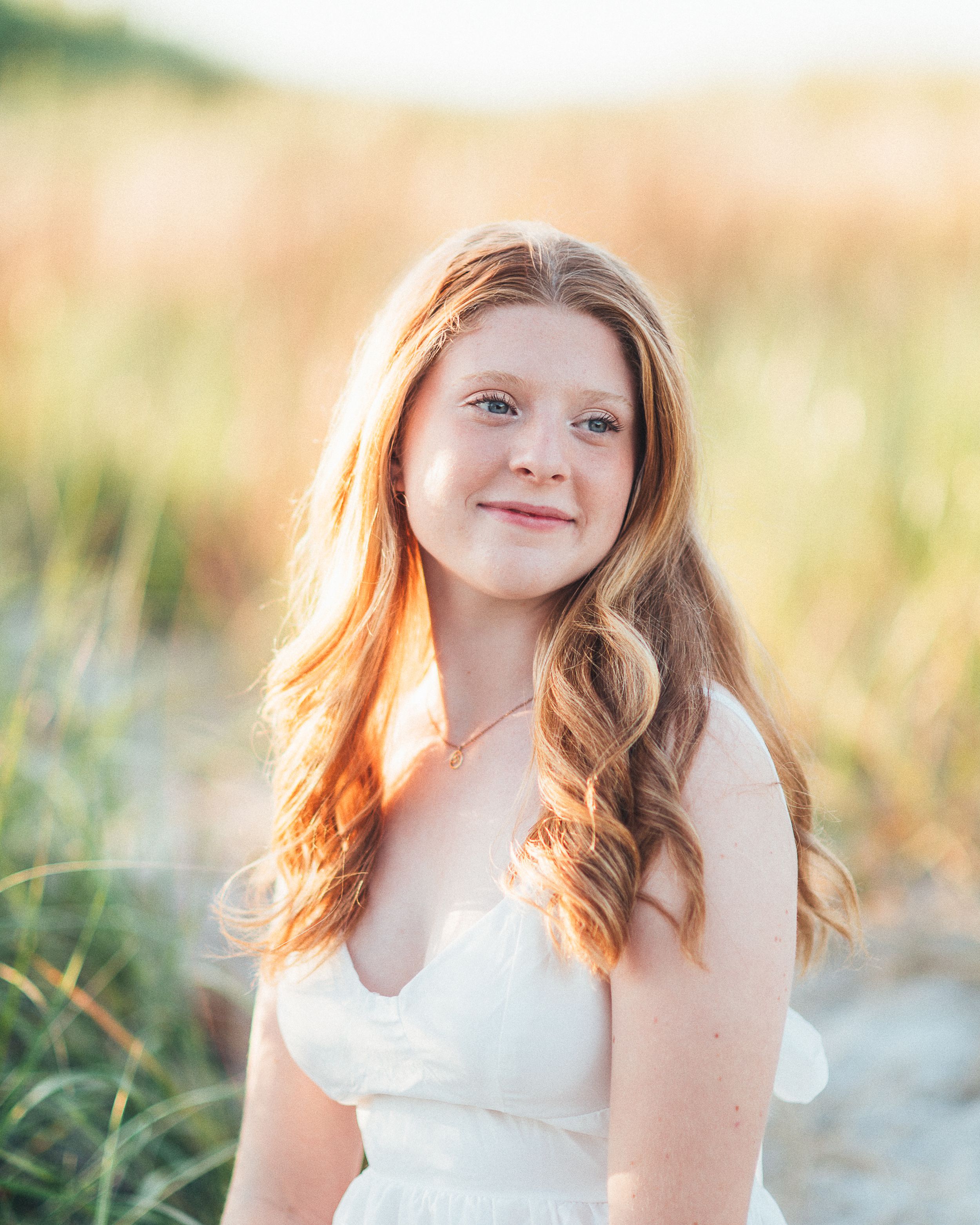 Young girl with long red hair in white dress smiling outdoors at golden hour near beach grass.