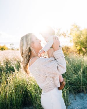 Person in a cream sweater holding and lifting up a baby against golden sunlight in a grassy coastal setting.