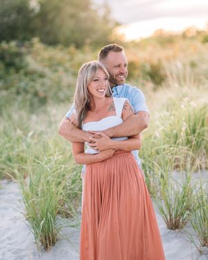 A couple laughs and embraces on a sandy beach path during golden hour as sunlight filters through coastal grasses.