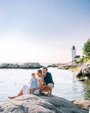 Couple lounging together on a rocky shoreline overlooking a scenic coastal lighthouse on a sunny summer day.