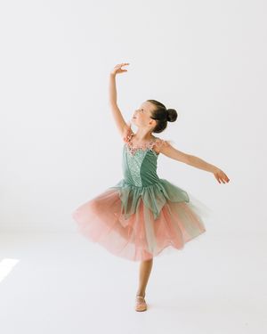 Young ballerina in green and pink tutu dress poses gracefully on pointe against white background.