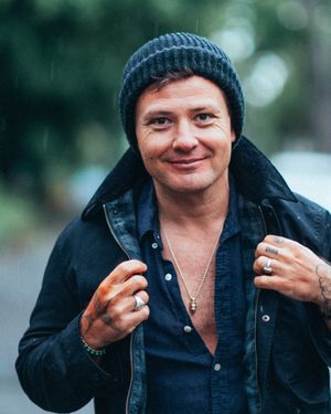 A smiling man wearing a blue beanie hat and dark jacket poses outdoors in light rain.