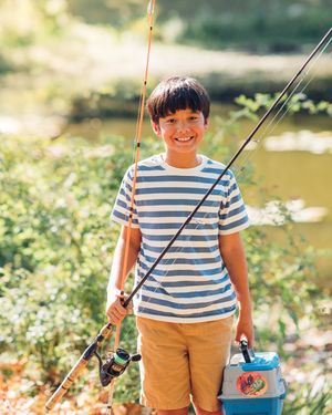 A child in a striped shirt and khaki shorts stands by a river with fishing gear on a sunny summer day.